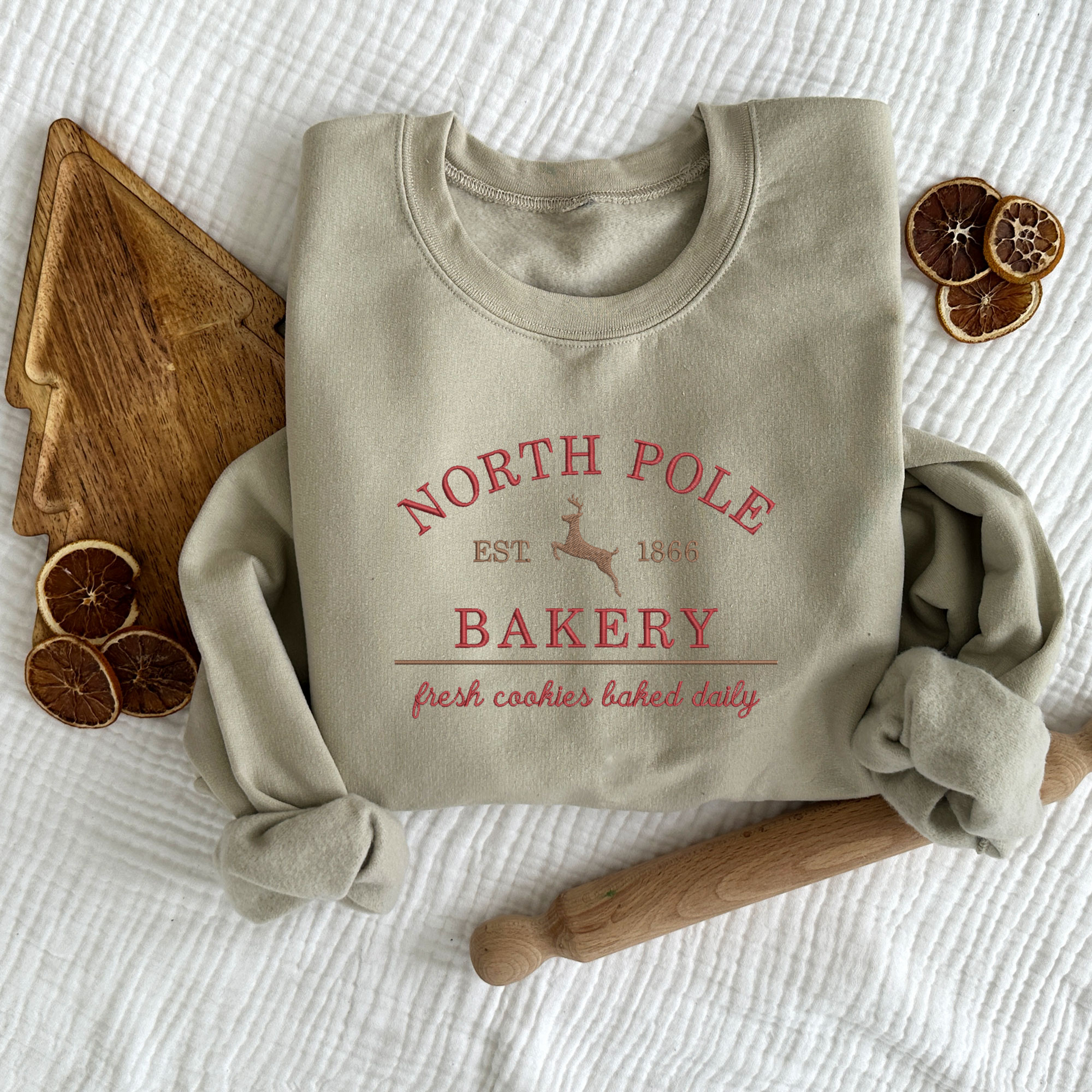 christmas styled flat lay of an ash crewneck sweatshirt with embroidered north pole bakery design in red and brown threads.