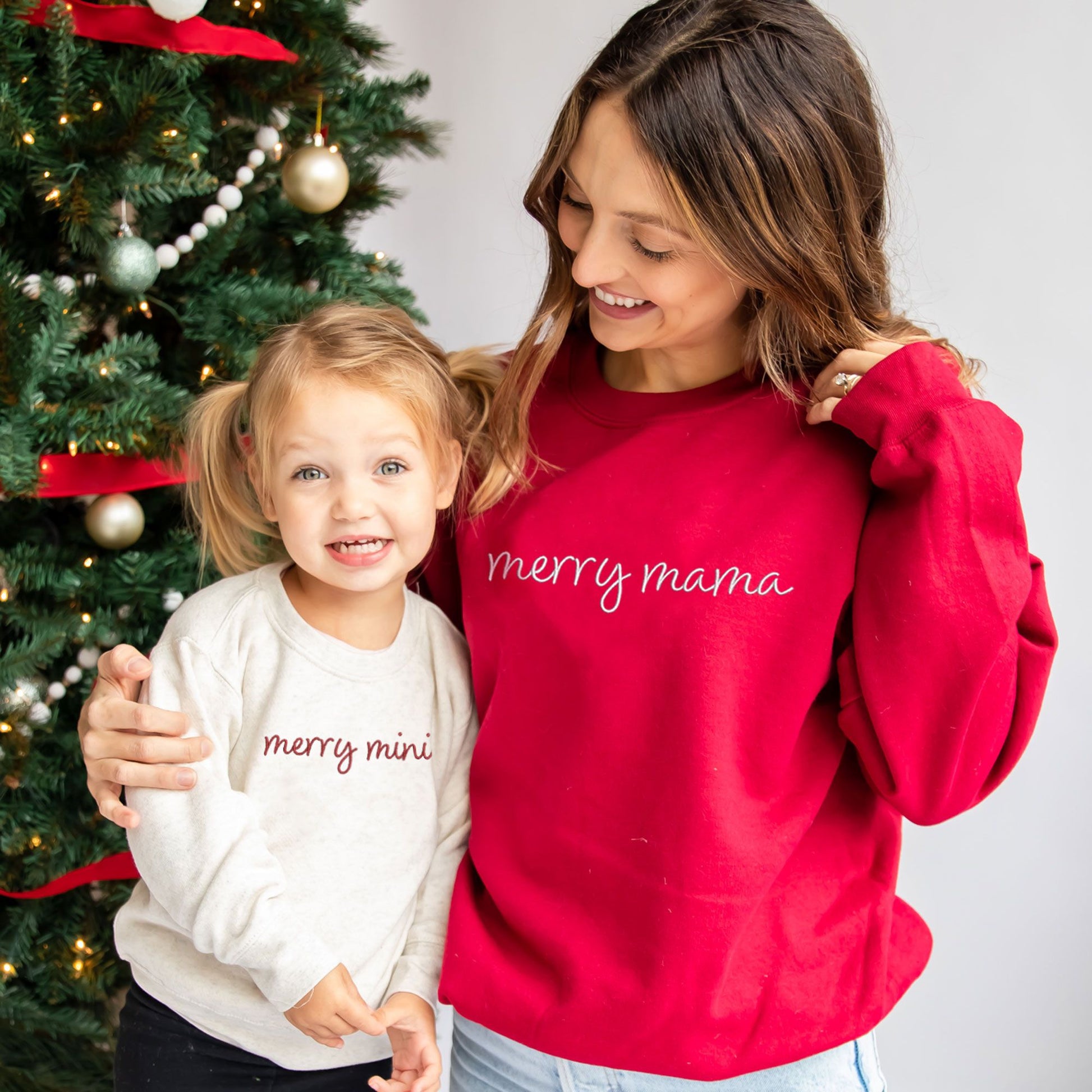 young mom and her daughter wearing matching Christmas crewneck sweatshirts. the toddler is wearing a heather oat crewneck with embroidered merry mini in red thread and the mom is wearing a red crewneck sweatshirt with merry mama in white thread.