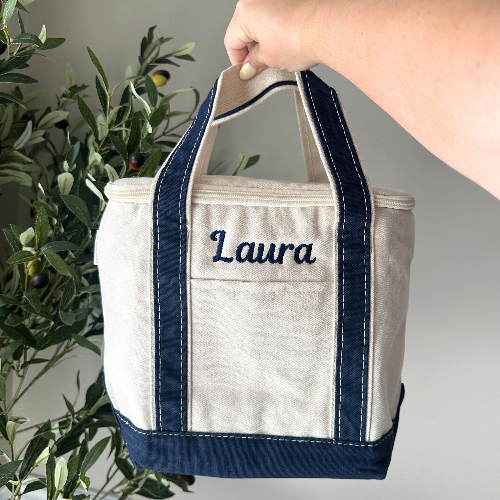 Beige tote bag with navy accents and 'Laura' embroidered on it, held by a hand against a neutral background.
