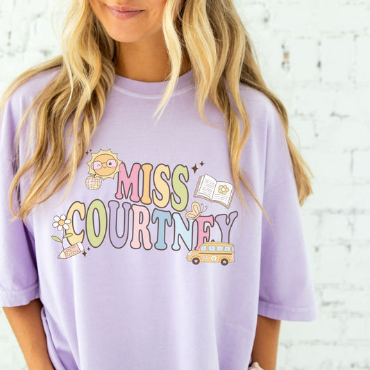 woman wearing a large orchid comfort colors t-shirt with custom name and fun pastel teacher icons printed on the front