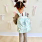 Child wearing a patterned backpack with 'SWE' monogrammed on it, standing against a wall with decorative ribbons.