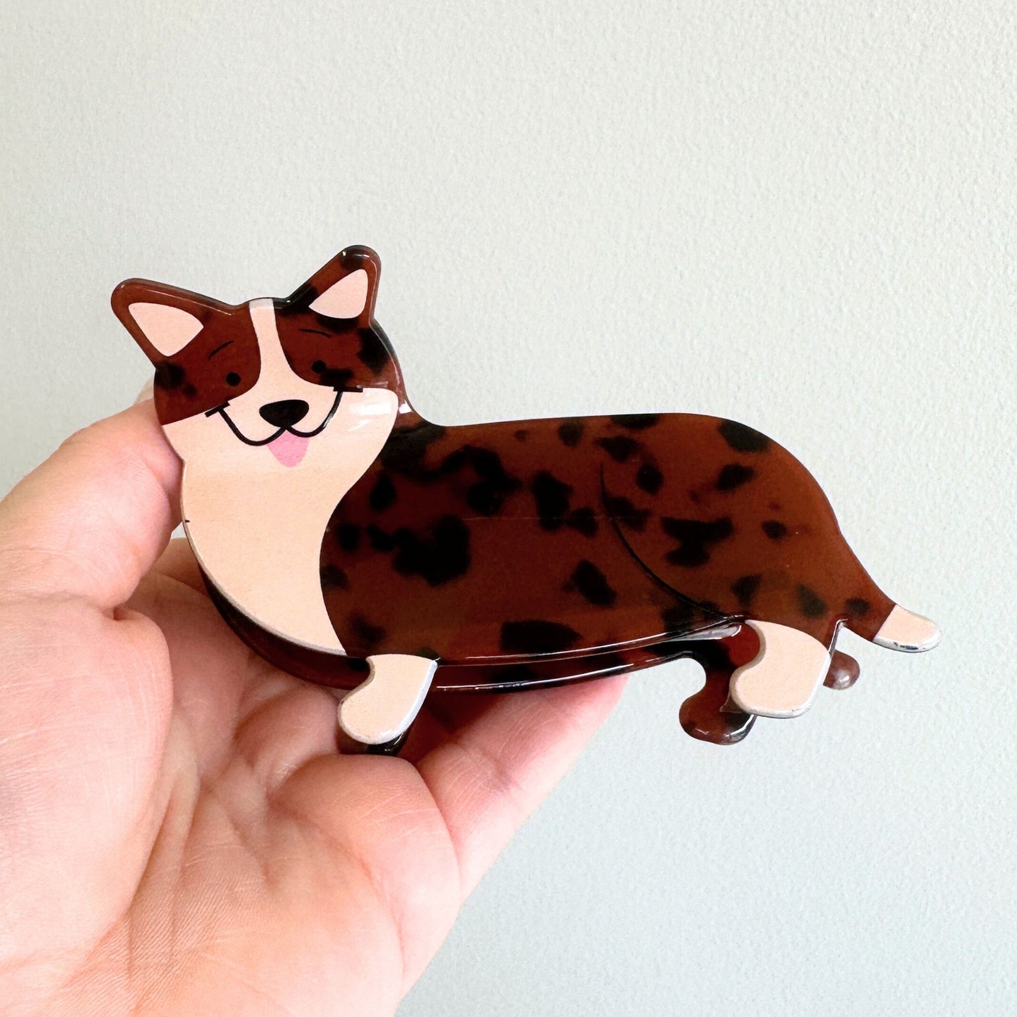 Hand holding a corgi-shaped hair clip with a brown and white pattern against a light background