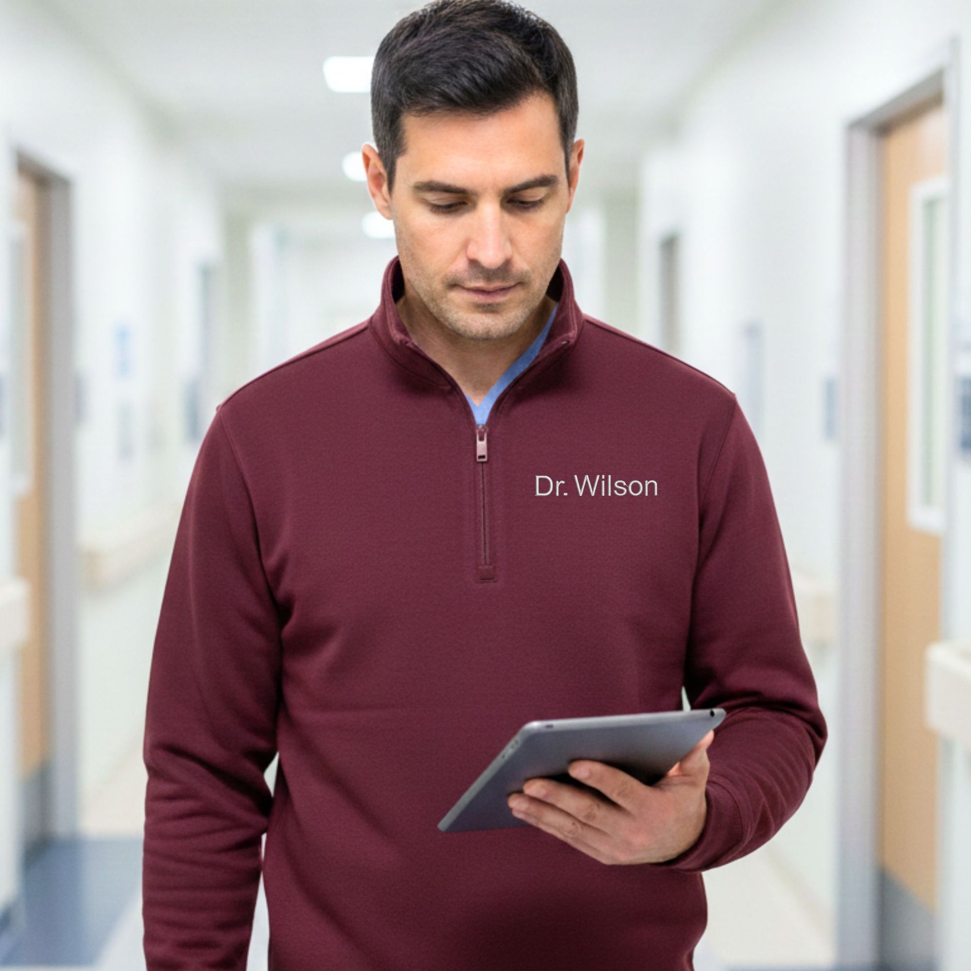 Man wearing a maroon pullover with 'Dr. Wilson' embroidered on it, holding a tablet in a hallway.