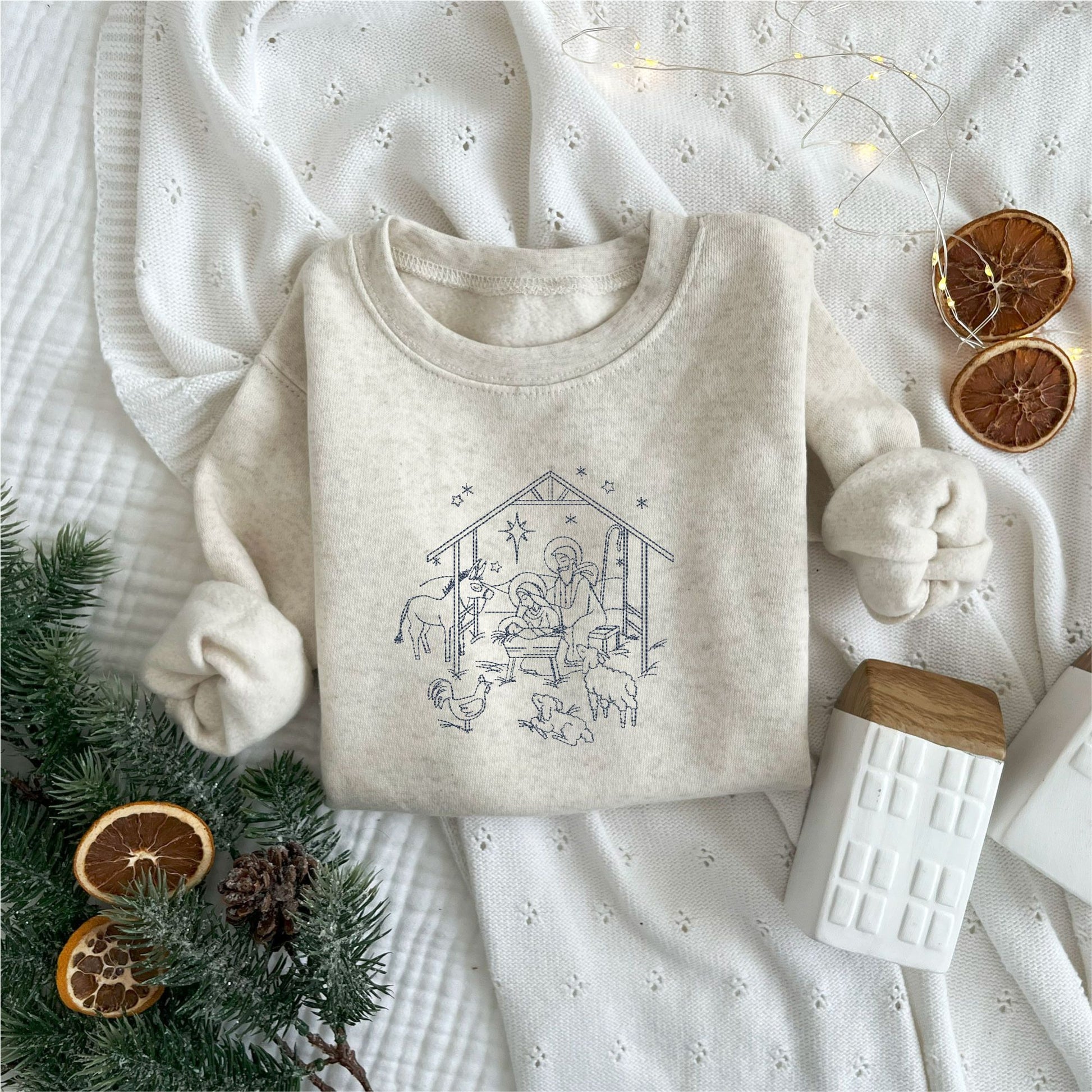 Christmas styled flat lay of a toddler heather oatmeal crewneck with embroidered stitched nativity scene in navy thread