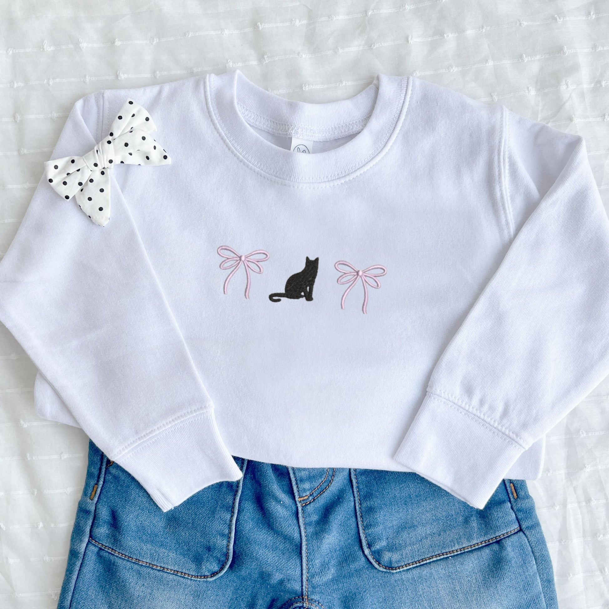 White youth sweatshirt with black cat and pink bows embroidered design, paired with blue jeans and a polka dot bow on a white background.