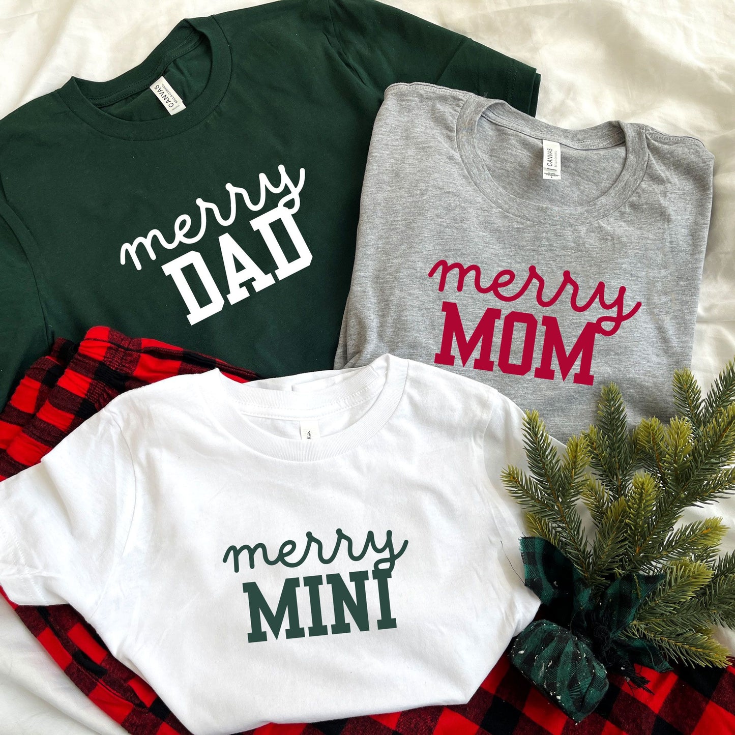 Photo of matching family Christmas pajama sets. Adult Forest green tee with merry dad printed in white ink, adult athletic heather tee with merry mom printed in red ink,  and youth white tee with merry mini printed in green ink.