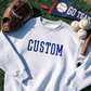 White sweatshirt with 'CUSTOM' text, baseball glove, baseballs, and 'GO TEAM!' banner on grass.