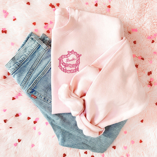 flat lay of jeans and a blush pink sweatshirt with a vintage heart cake embroidered design on the left chest with custom text reading 'books' centered on the top of the cake in pink thread 