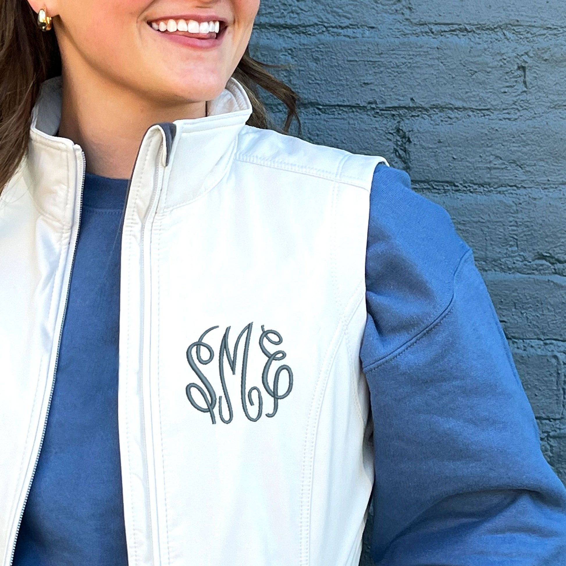 young woman wearing a white softshell vest with embroidered monogram on the left chest in french blue thread.