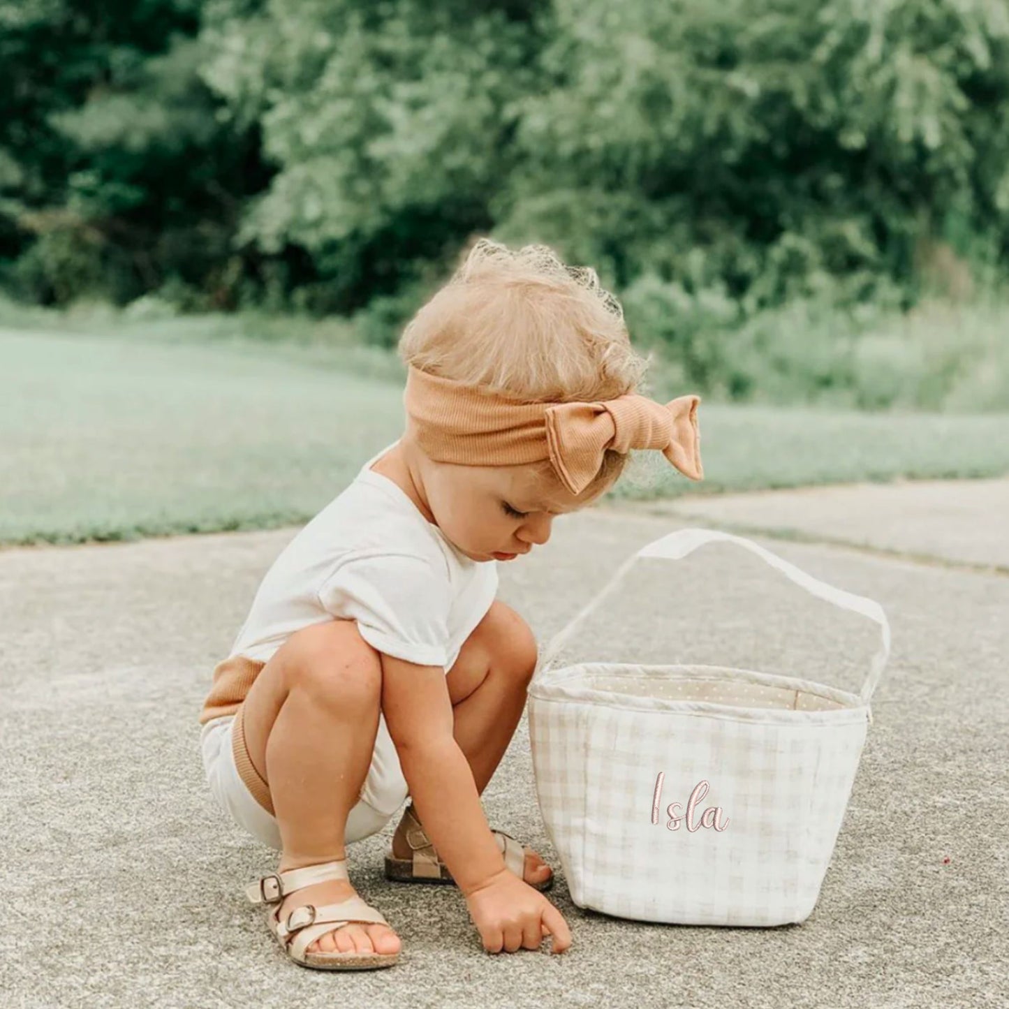 Child sitting on the ground next to a tan gingham easter basket with 'Isla' written on it, outdoors.