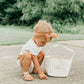 Child sitting on the ground next to a tan gingham easter basket with 'Isla' written on it, outdoors.