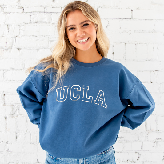 young woman wearing an indigo crewneck sweatshirt with custom athletic block outline embroidered font across the chest that spells out UCLA