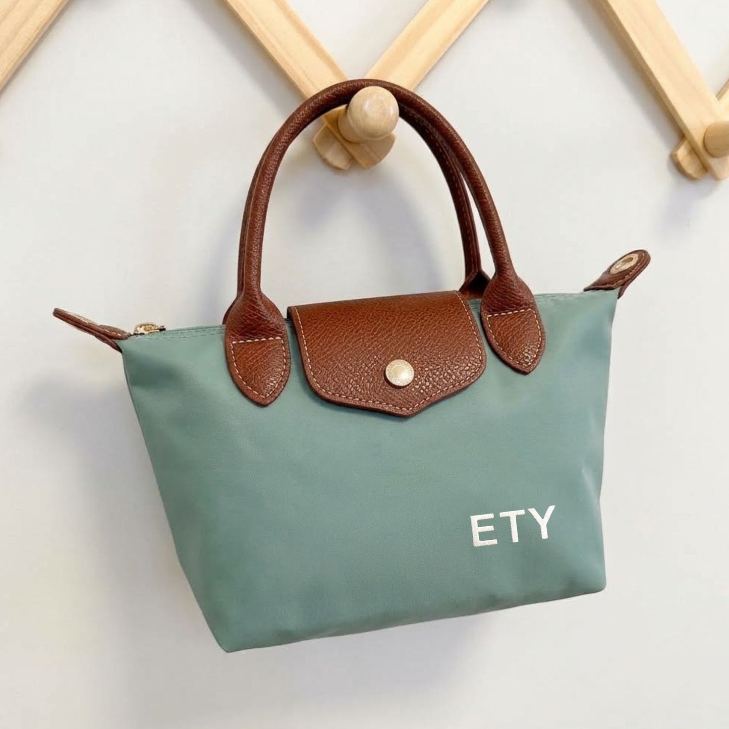 A small structured handbag in muted sage green with brown leather handles and flap, hanging on a wooden display stand against a light wall. The flap has a gold-tone snap, and the bag is embroidered with the white letters 'ETY'.