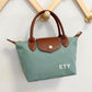 A small structured handbag in muted sage green with brown leather handles and flap, hanging on a wooden display stand against a light wall. The flap has a gold-tone snap, and the bag is embroidered with the white letters 'ETY'.