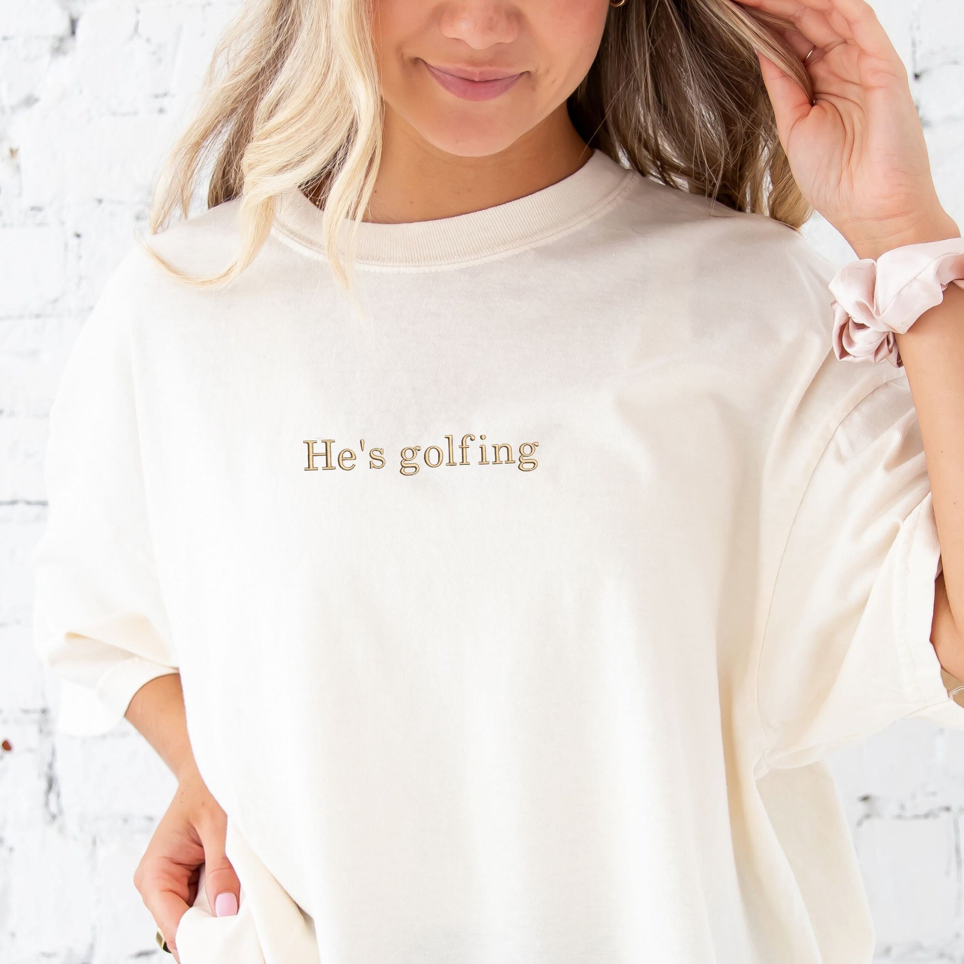 woman wearing an ivory comfort colors t-shirt with custom he's golfing embroidered design in camel thread cross the chest 