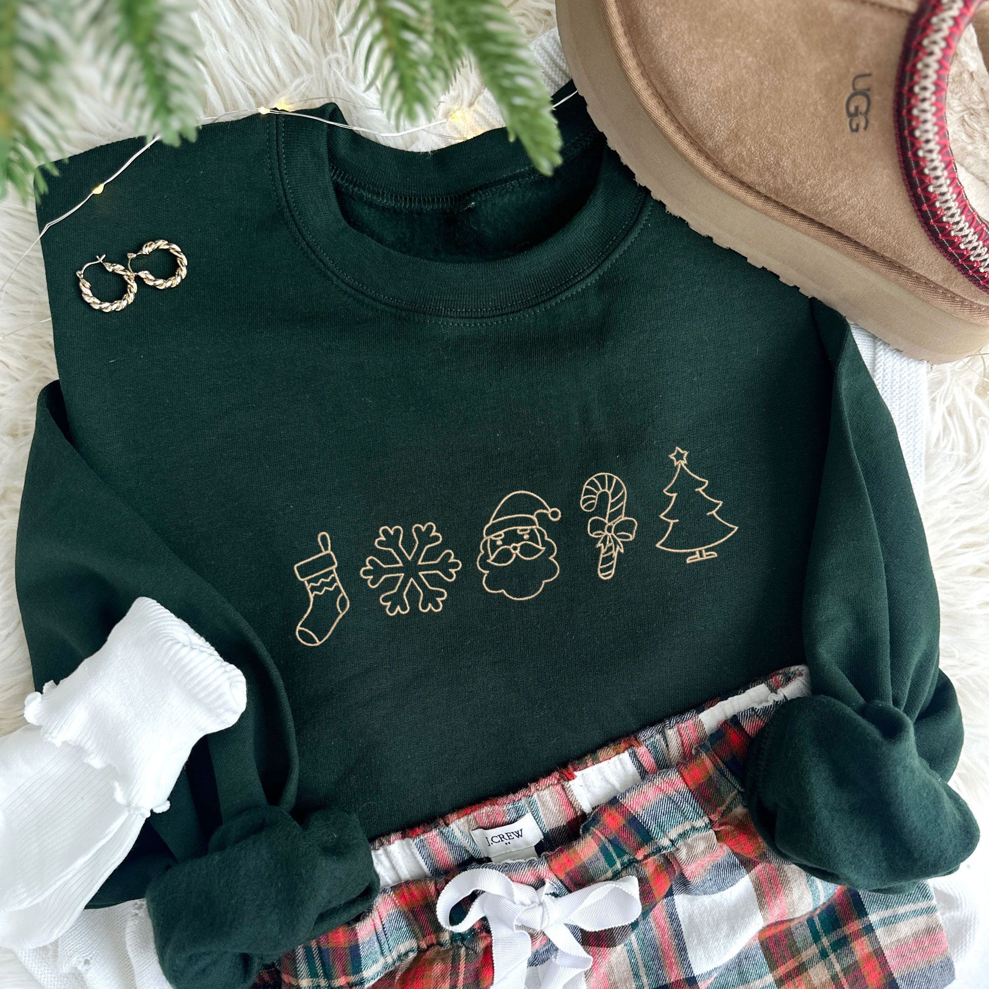 Christmas styled flat lay photo of a forest green crewneck sweatshirt with Christmas outlined icons embroidered across the chest in camel thread.