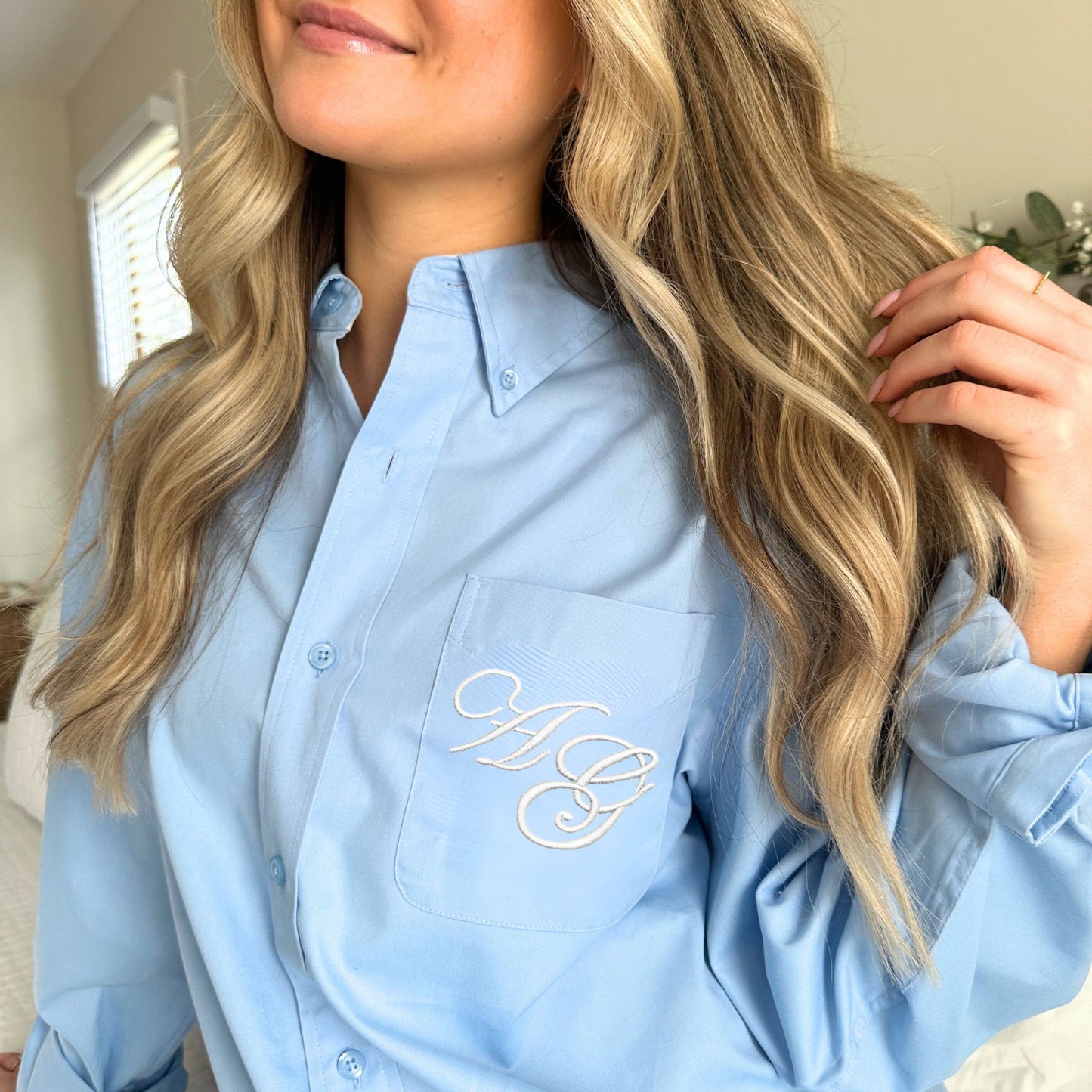 Woman wearing a blue button up shirt with monogrammed Edwardian script initials on the pocket  in a home setting