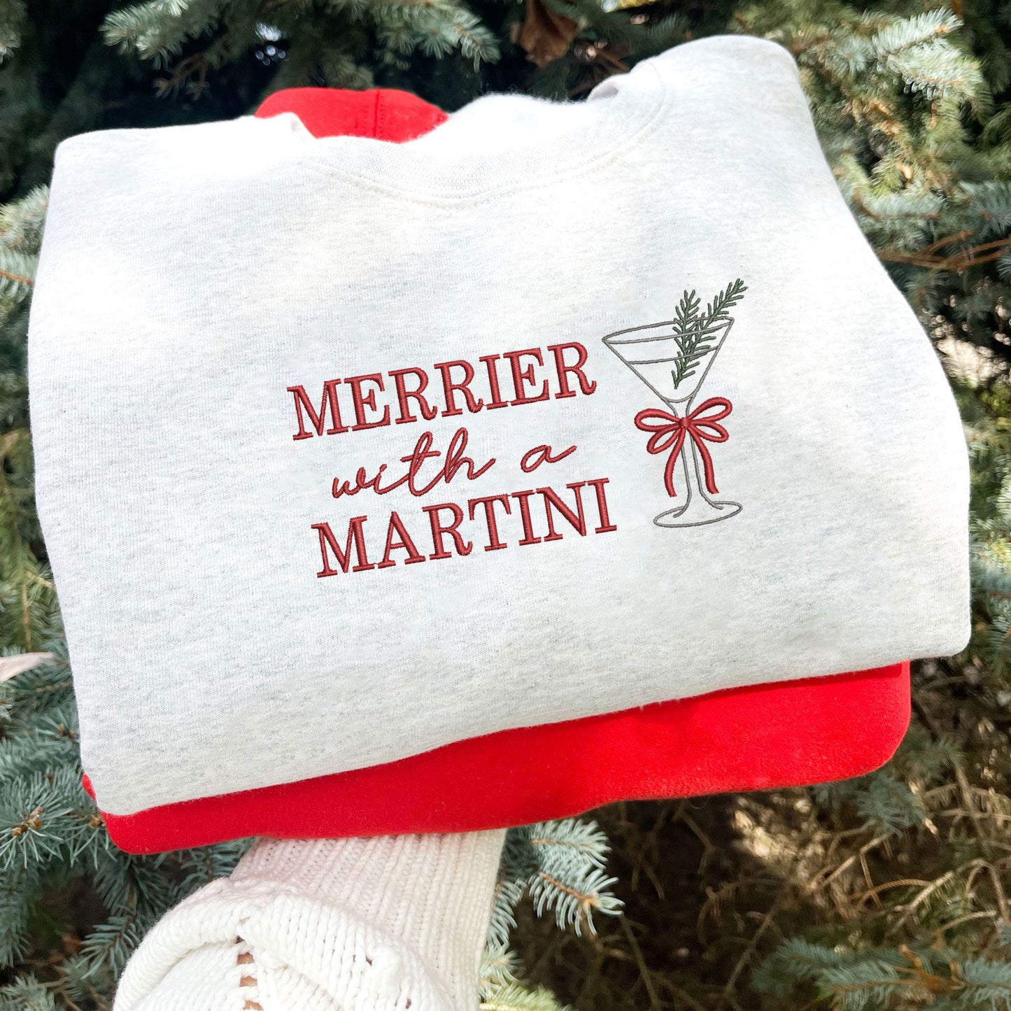 ash crewneck sweatshirt with embroidered merrier with a martini text in red thread and martini glass design with bow and pine sprig across the chest