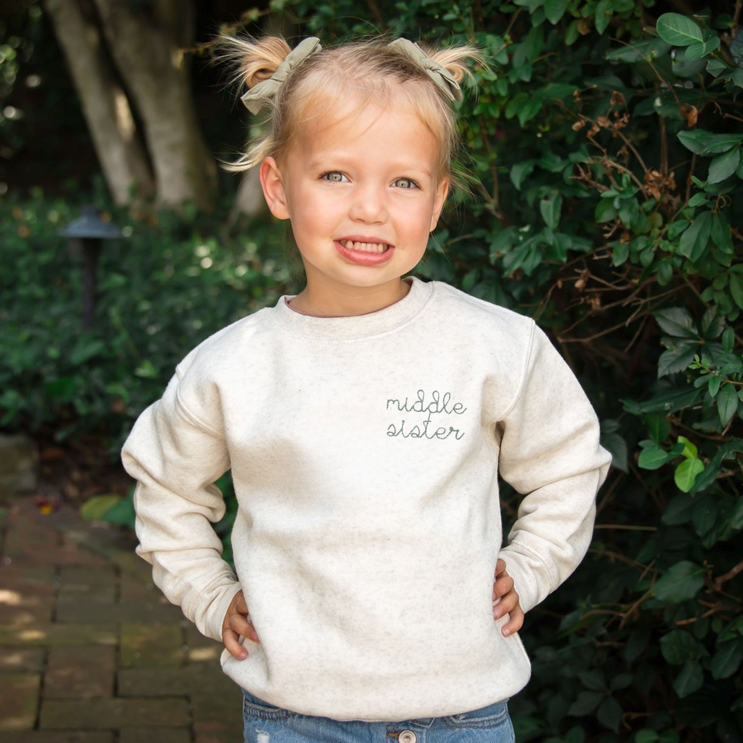 little girl wearing a natural heather crewneck with custom middle sister embroidered design on the left chest in eucalyptus thread