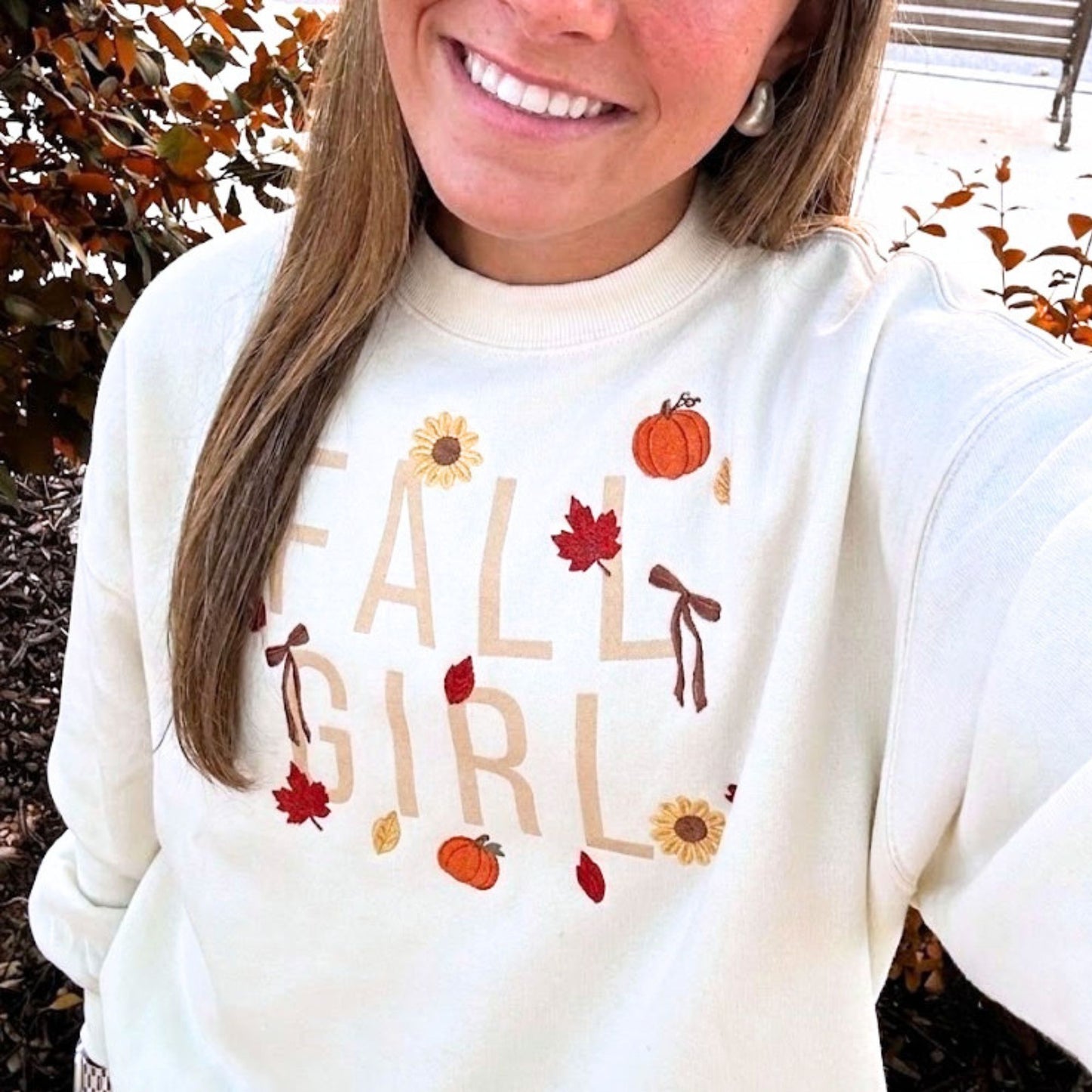 A woman wearing a cream-colored crewneck sweatshirt with a fall-themed graphic that reads 'FALL GIRL' in beige letters, decorated with orange pumpkins, red and yellow leaves, and brown bows.