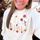 A woman wearing a cream-colored crewneck sweatshirt with a fall-themed graphic that reads 'FALL GIRL' in beige letters, decorated with orange pumpkins, red and yellow leaves, and brown bows.
