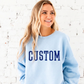 Woman wearing a light blue sweatshirt with 'CUSTOM' printed on it against a white brick wall.