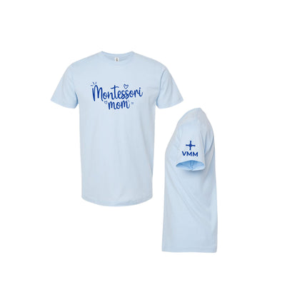 A light blue short-sleeve t-shirt shown from the front and side. The front features the text 'Montessori mom' in blue script, and the side view shows 'VMM' with a small cross printed on the sleeve.