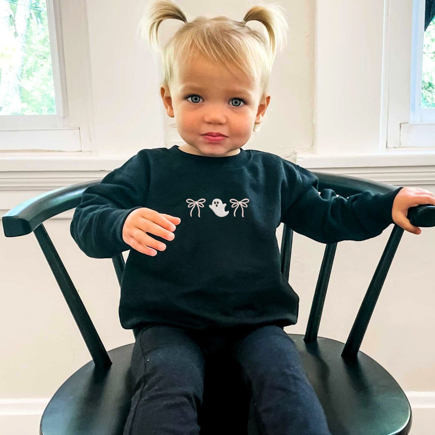 Child wearing a black sweatshirt with an embroidered double loop bow and ghost design. she's sitting on a black chair.