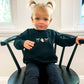Child wearing a black sweatshirt with an embroidered double loop bow and ghost design. she's sitting on a black chair.