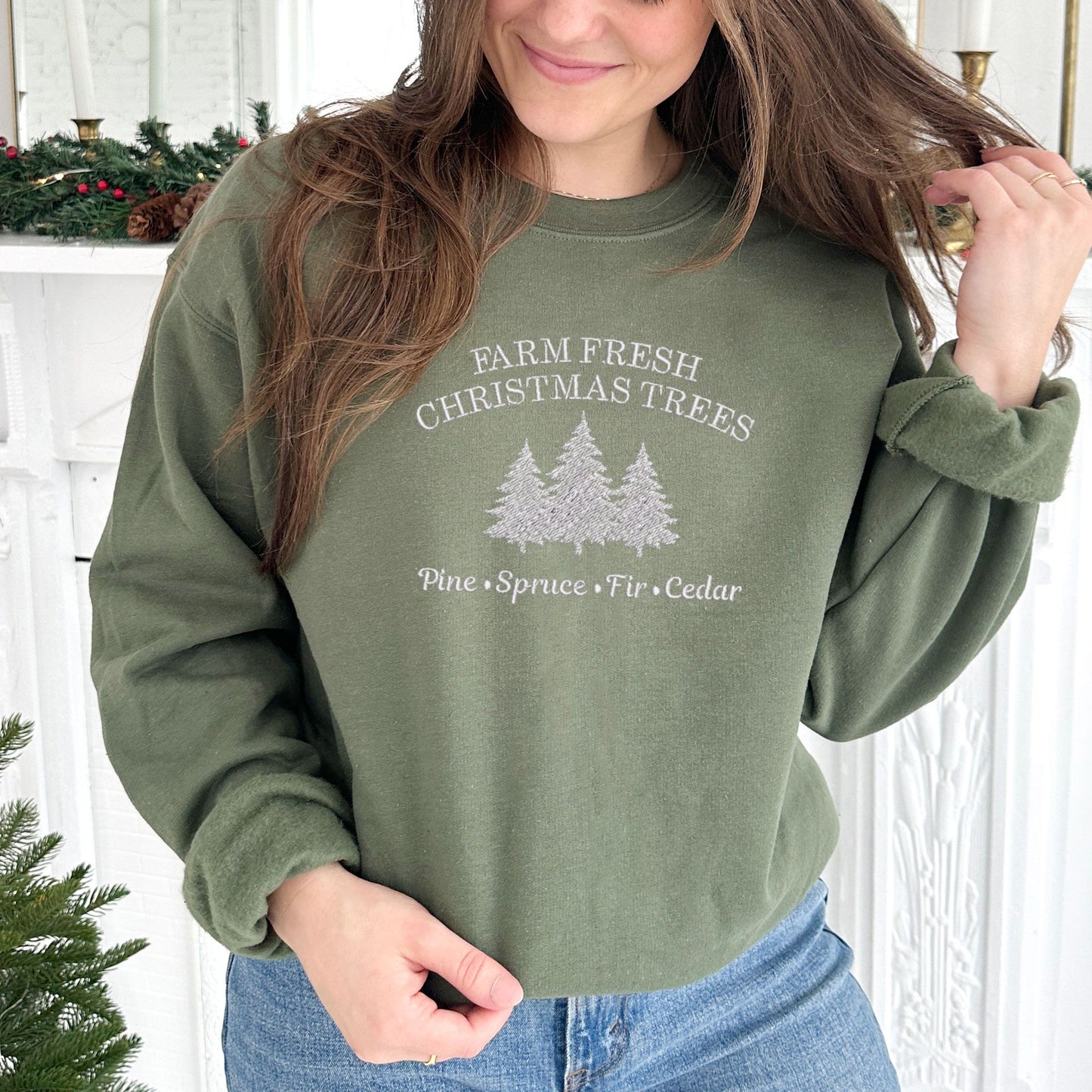 young woman wearing a military green crewneck sweatshirt with embroidered farm fresh Christmas trees design with Christmas tree embroidery across the chest in white thread