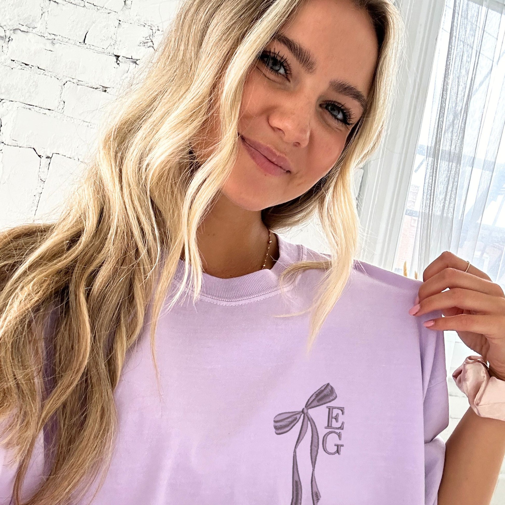 girl wearing an orchid comfort colors t-shirt with custom ribbon bow stacked initial embroidered design on the left chest in smoky orchid thread.