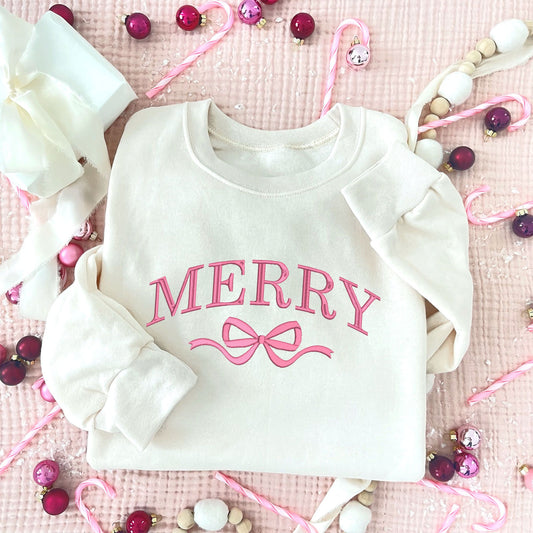 Christmas styled flat lay of a sweet cream crewneck with embroidered merry and bow design in pink thread