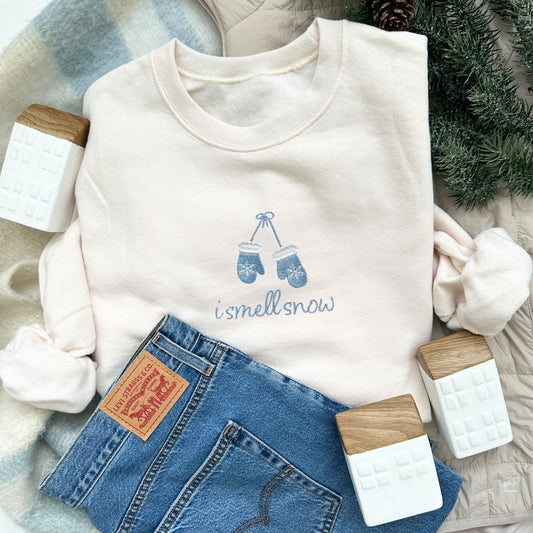 sweet cream crewneck sweatshirt with i smell snow and mittens embroidered on the center chest in powder blue thread