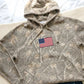 Camouflage hoodie with an American flag patch on a neutral background
