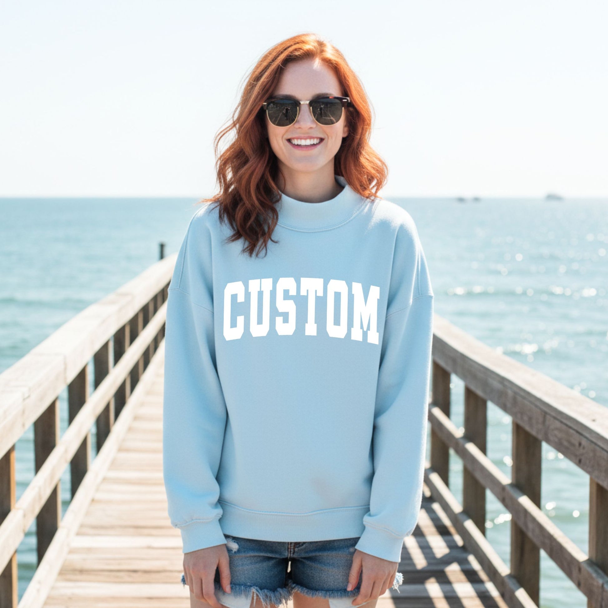Person wearing a light blue sweatshirt with 'CUSTOM' text on a wooden pier by the water.