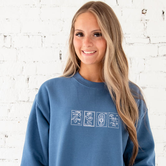 a girl standing in front of a white brick wall wearing an indigo sweatshirt with 4 embroidered Italian inspired stamps across the chest in white thread. The stamps feature lemons, a lobster, a beach scene, and seashells.