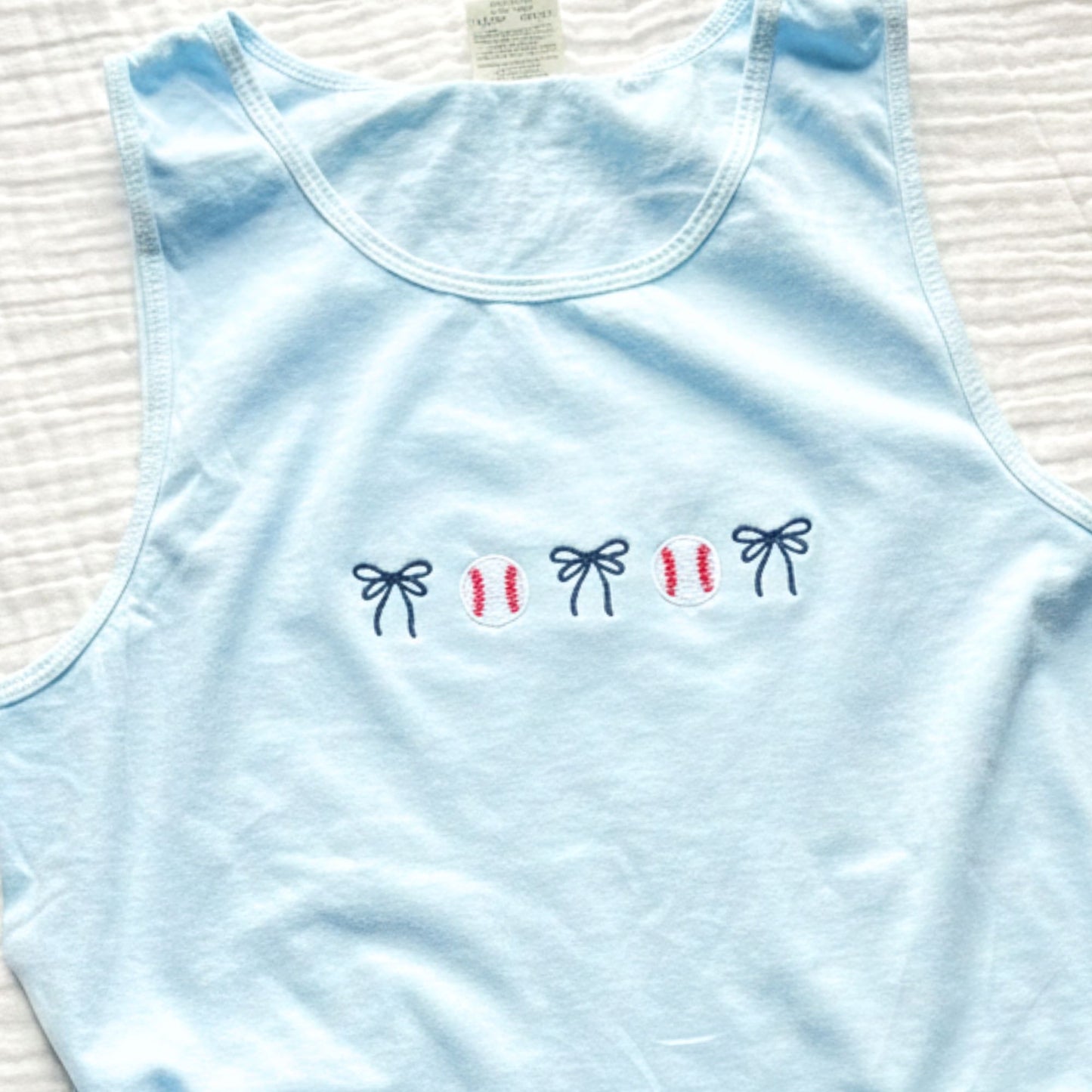 Light blue tank top with embroidered baseball and bow icons on a textured surface
