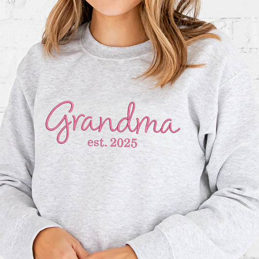 close up of a woman wearing an ash  crewneck sweatshirt with grandma est 2025 embroidered across the chest in pink thread