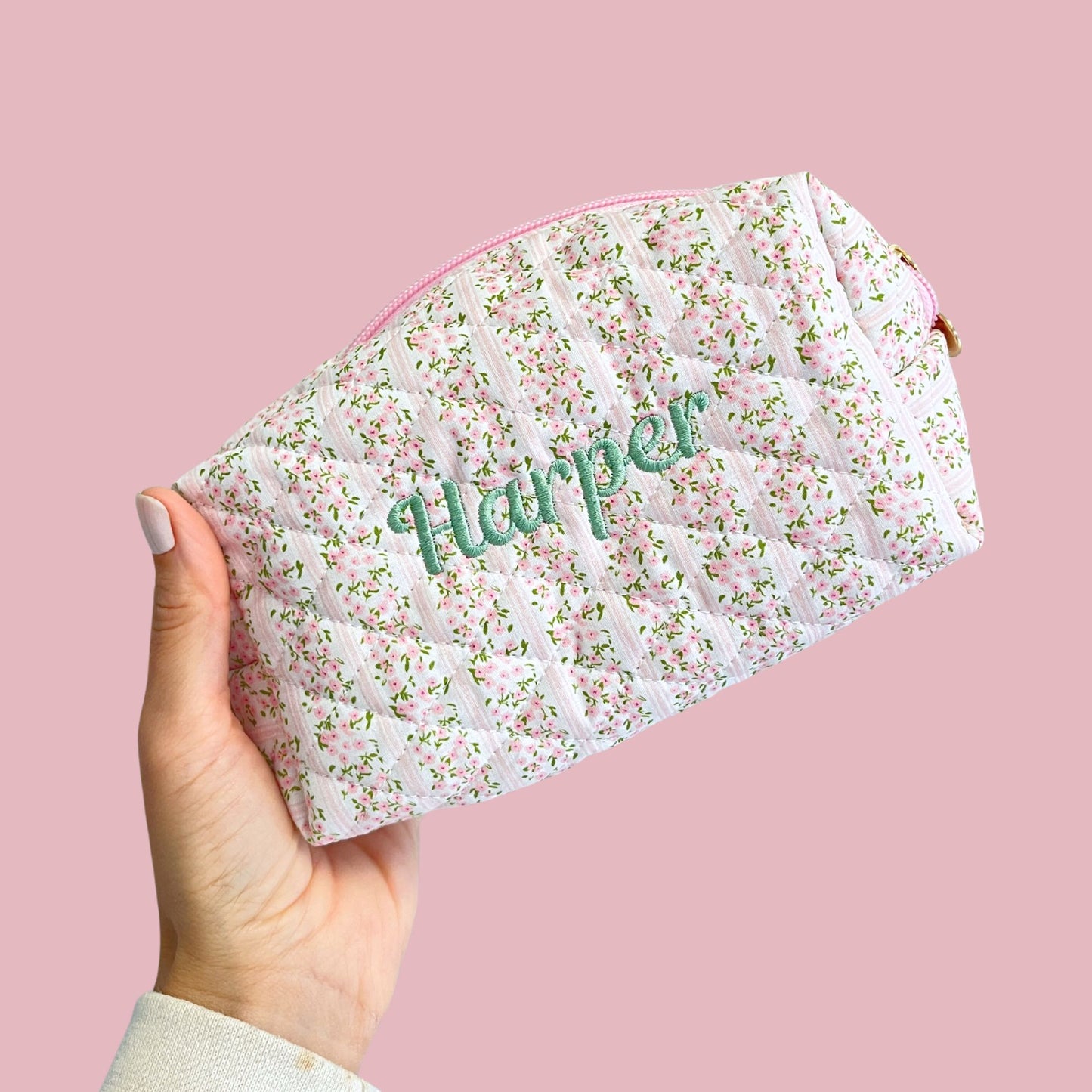 Floral-patterned pouch with 'Harper' embroidered on it held against a pink background