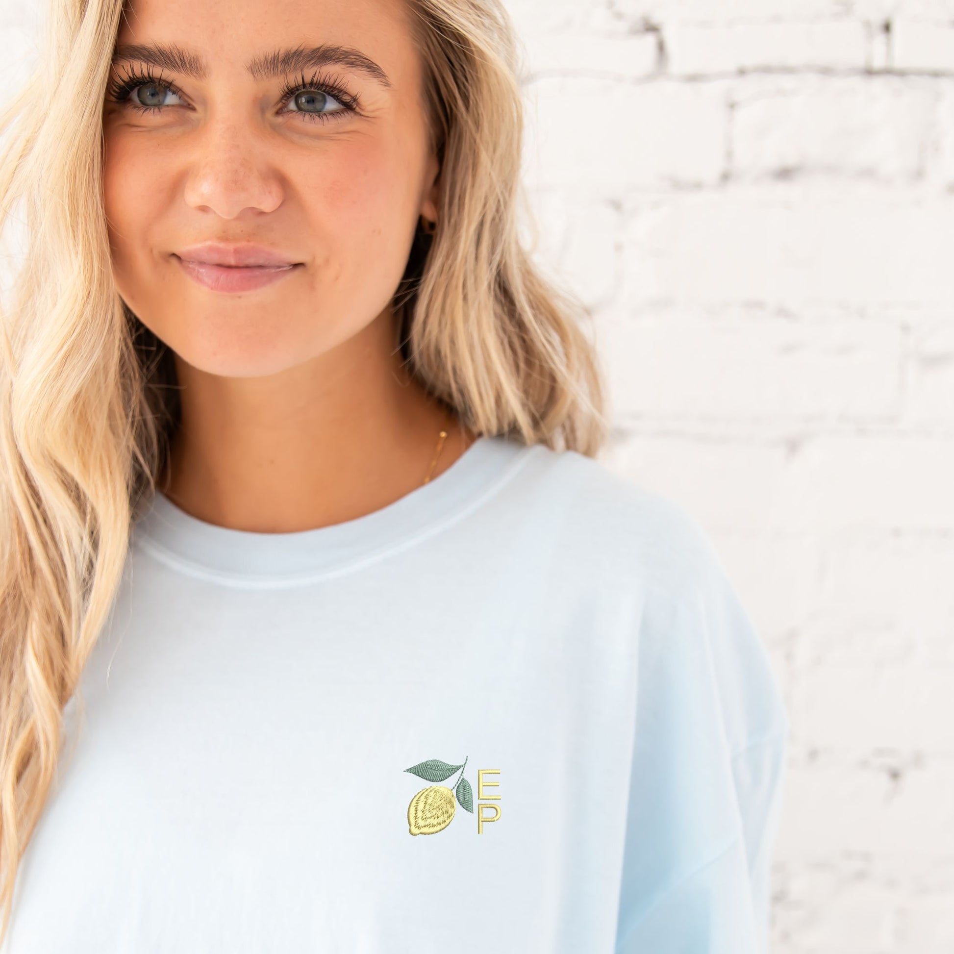 Girl standing in front of a white brick wall wearing an oversized chambray comfort colors t-shirt with a custom embroidered design on the left chest featuring a mini lemon and 2 initials