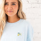 Girl standing in front of a white brick wall wearing an oversized chambray comfort colors t-shirt with a custom embroidered design on the left chest featuring a mini lemon and 2 initials