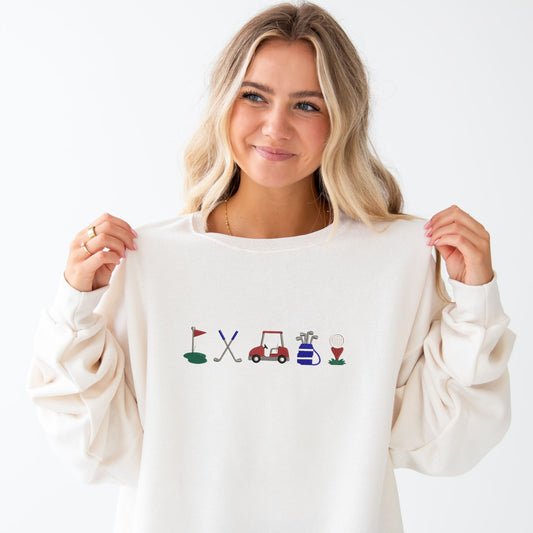 woman wearing a sweet cream crewneck sweatshirt with custom embroidered golf icons - golf flag, golf poles, golf cart, golf bag, and ball with tee in shades of blue, green, and red