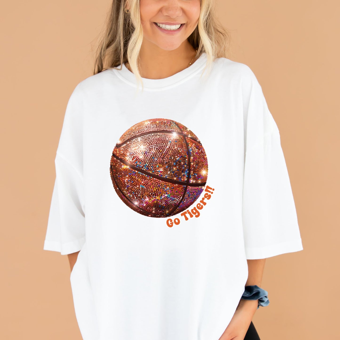 young woman wearing a white comfort colors t-shirt with large glitter basketball print with Go tigers text wrapping around the ball in orange ink