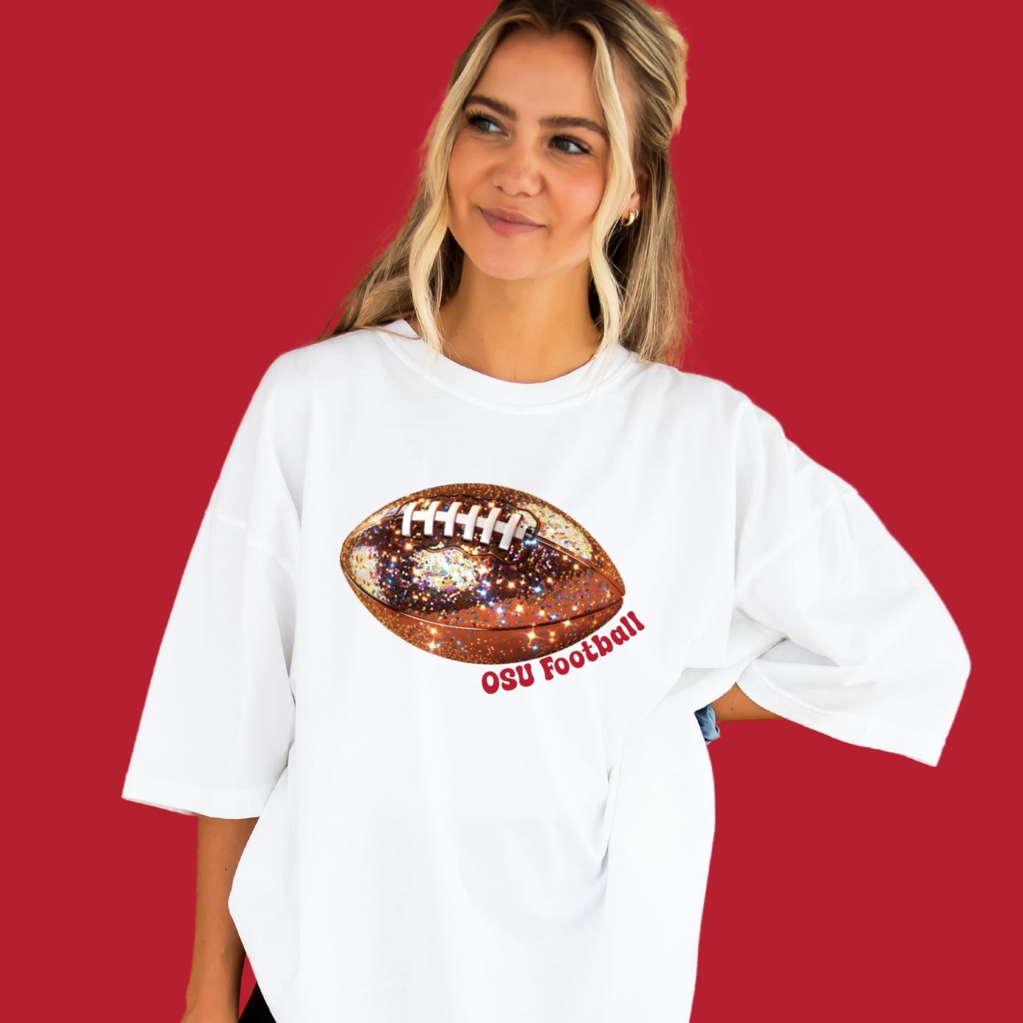 young woman wearing a white comfort colors tee with large glitter football print and osu football text curved under the football in red ink