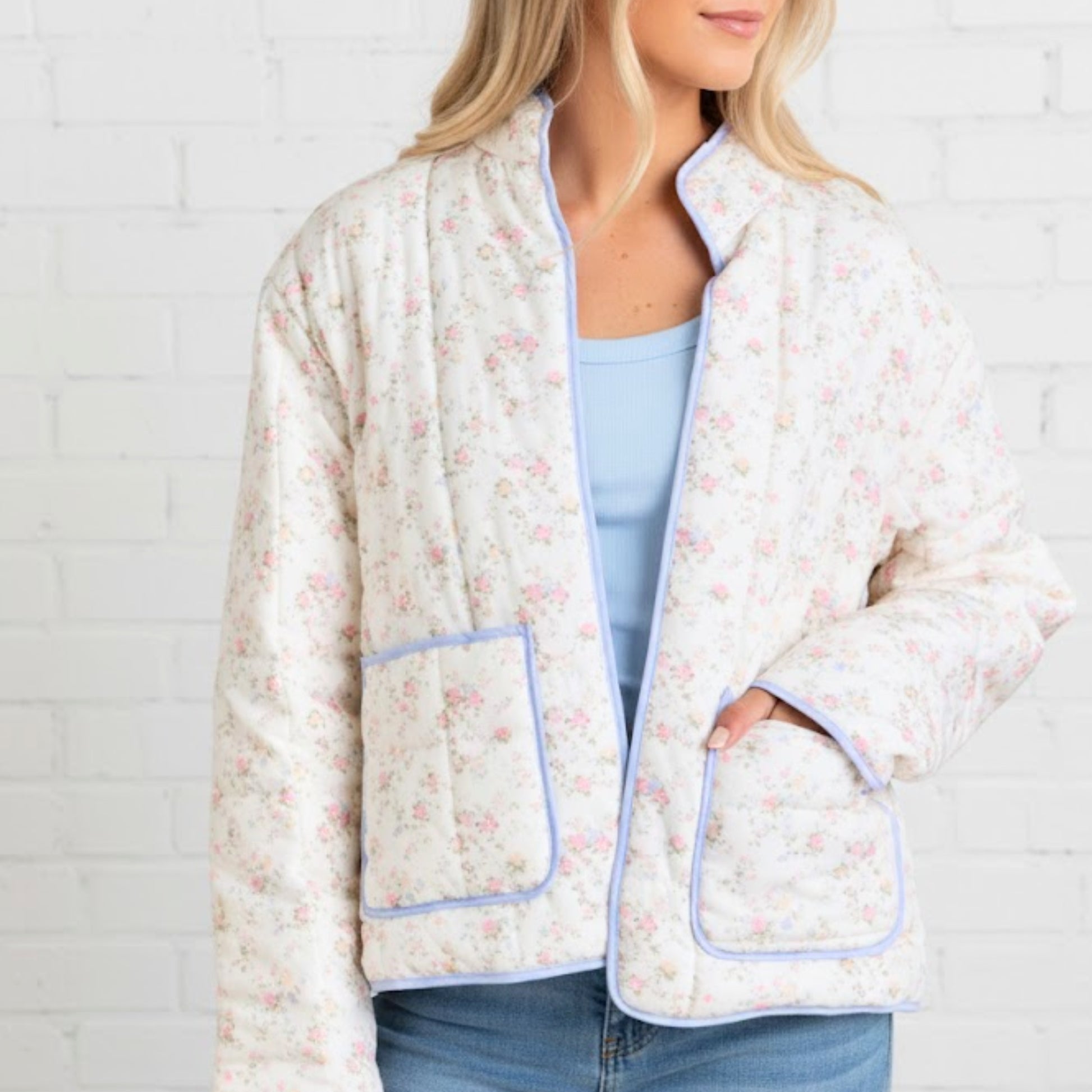 Woman wearing a floral patterned jacket with blue trim against a white brick wall.