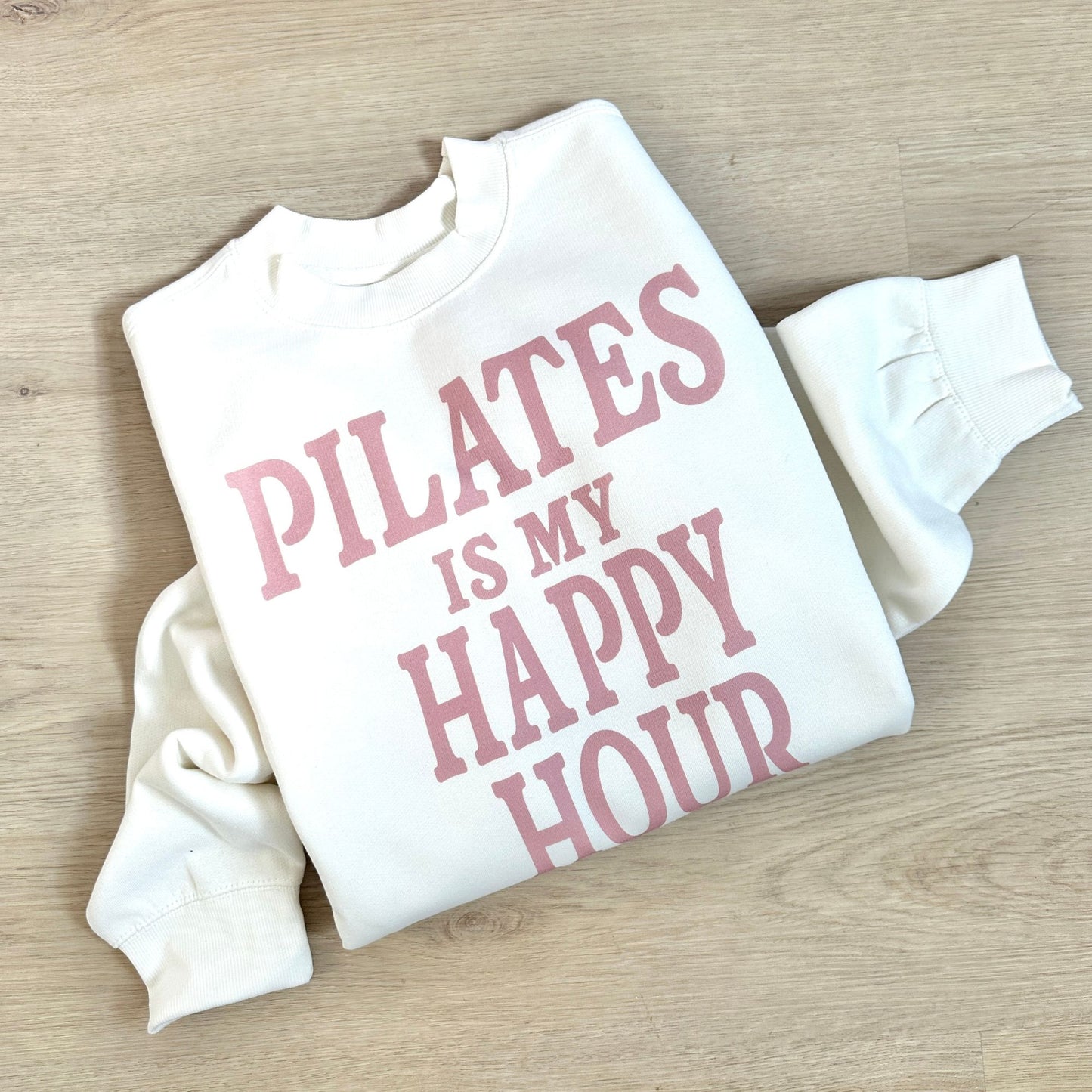 White sweatshirt with pink text 'PILATES IS MY HAPPY HOUR' on a wooden surface