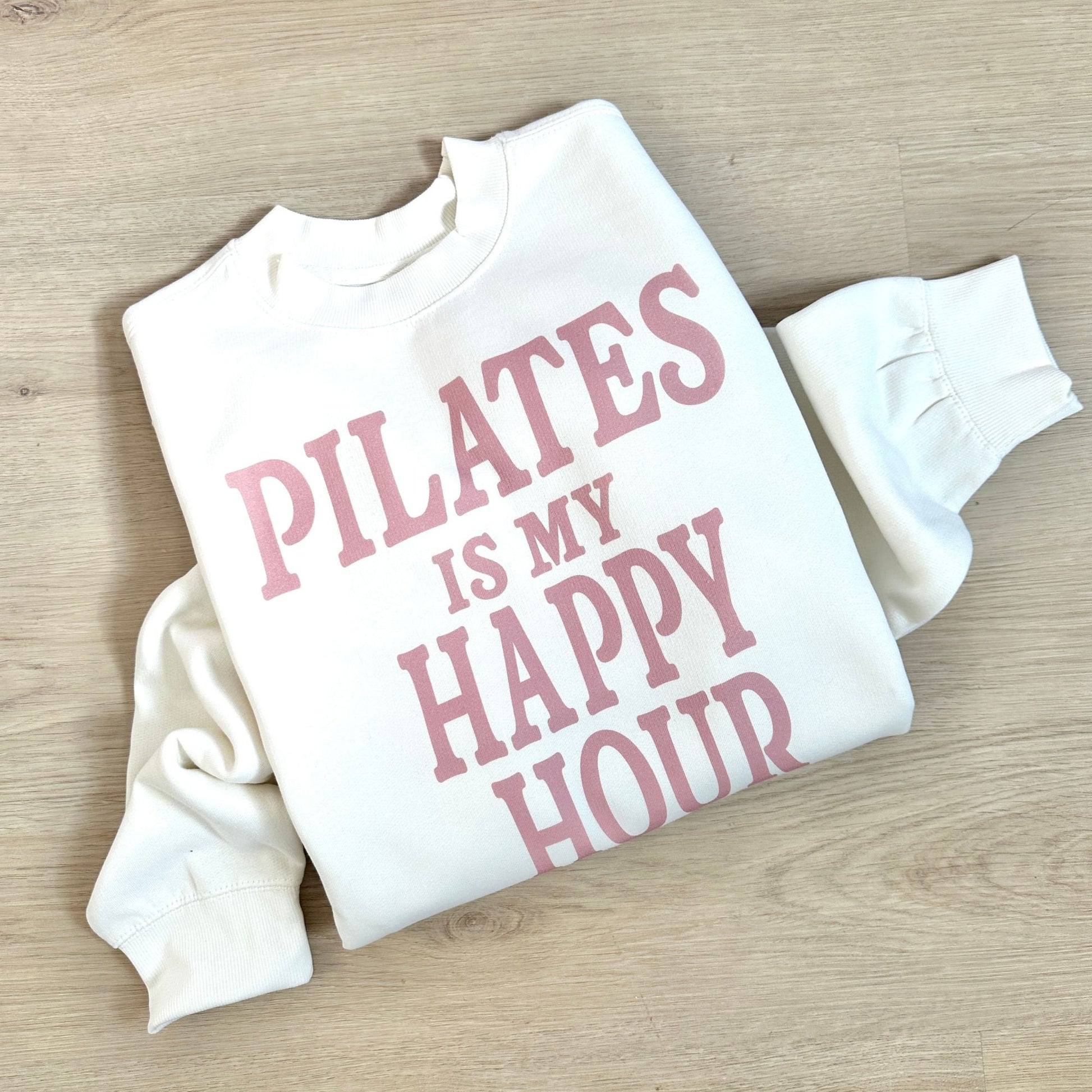 White sweatshirt with pink text 'PILATES IS MY HAPPY HOUR' on a wooden surface