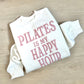 White sweatshirt with pink text 'PILATES IS MY HAPPY HOUR' on a wooden surface