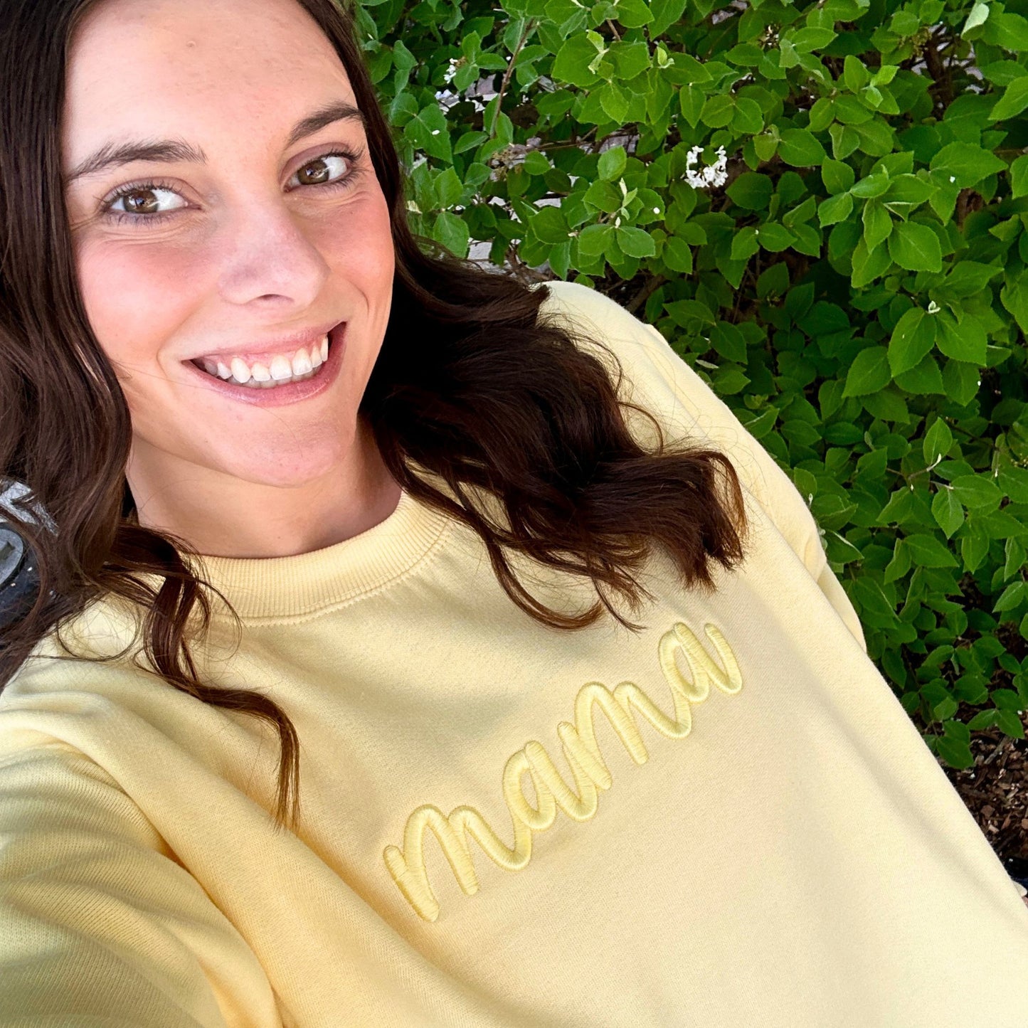 Woman wearing a yellow sweatshirt with 'mama' on it, standing in front of green foliage.
