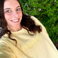 Woman wearing a yellow sweatshirt with 'mama' on it, standing in front of green foliage.